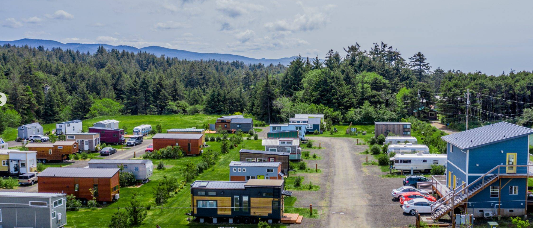 Aerial view of Tiny Tranquility Park in Oregon showing rows of modern tiny homes on wheels and vintage trailers surrounded by pine trees, grassy fields, and distant mountains under a partly cloudy sky.