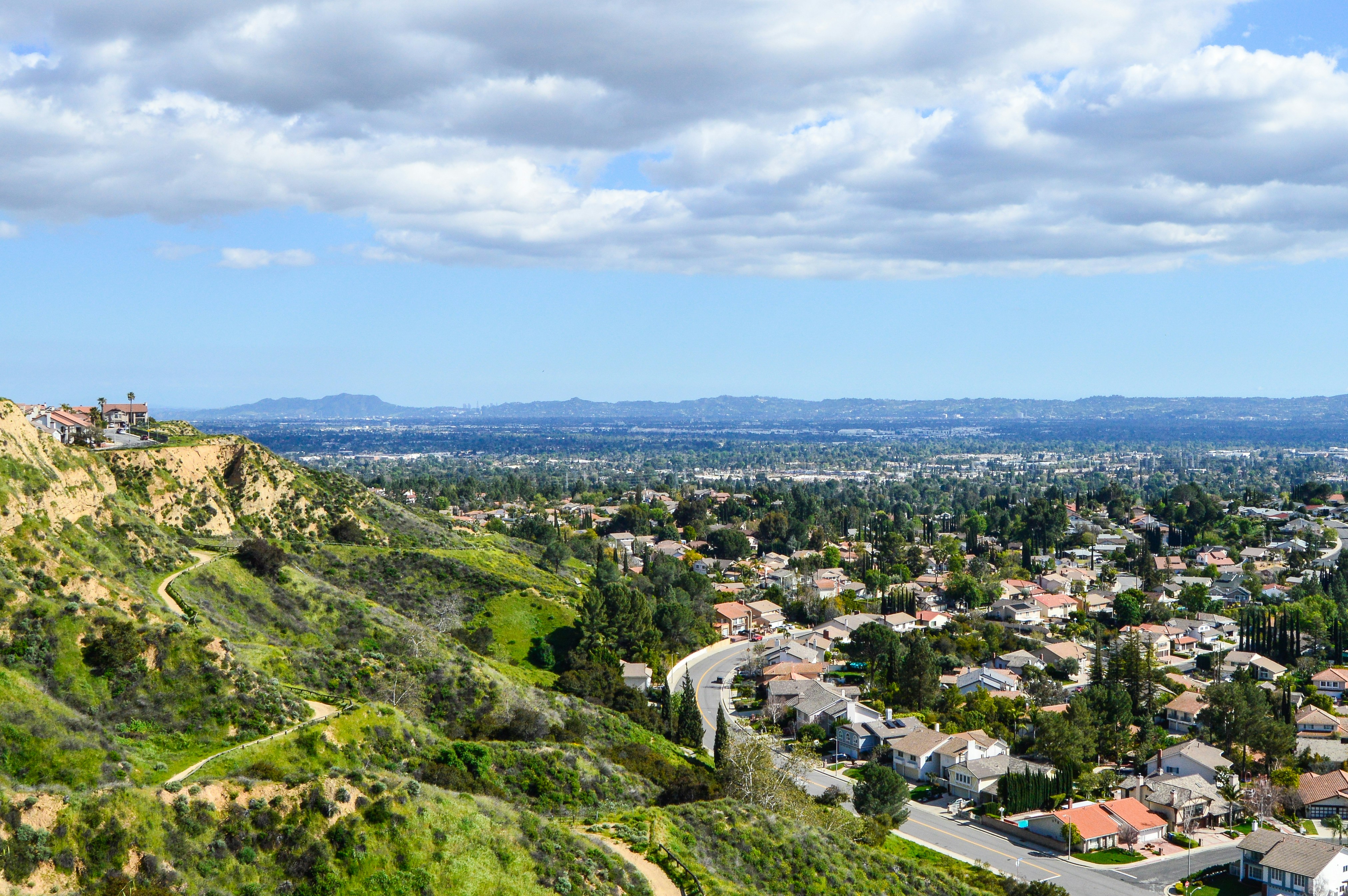 Aerial view of Los Angeles hillside neighborhoods with homes, greenery, and skyline in the distance under a clear California sky.