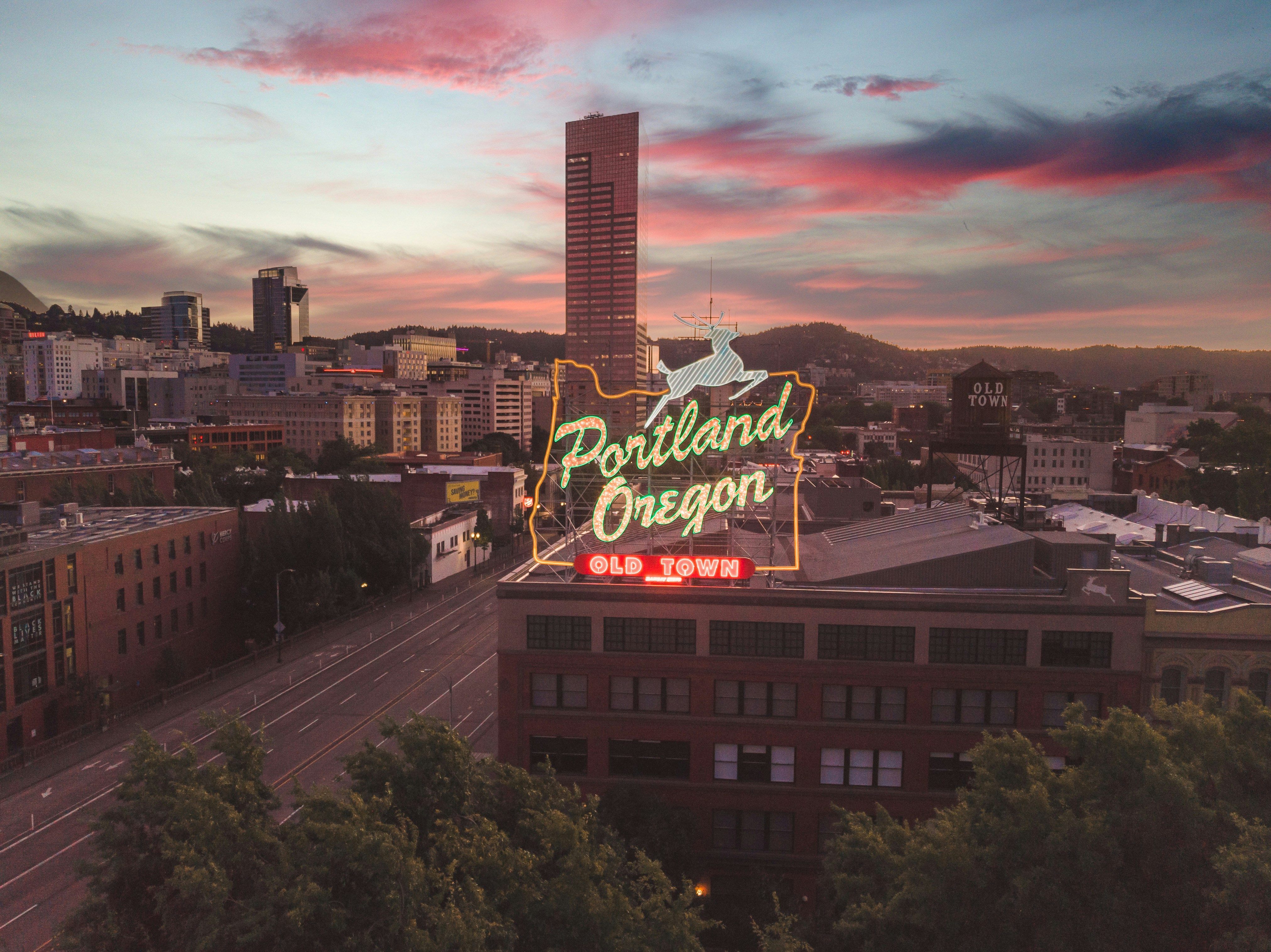 Aerial view of downtown Portland, Oregon at sunset with the illuminated “Portland Oregon Old Town” sign in the foreground, surrounded by city buildings and hills under a colorful sky.