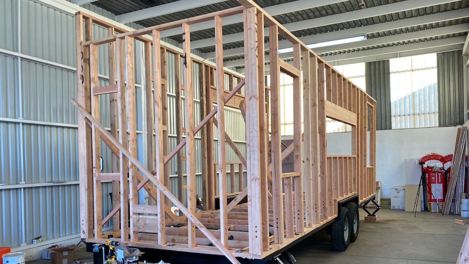 A partially constructed tiny home frame sits on a trailer inside a workshop. The wooden frame outlines walls and windows, with diagonal bracing for structural support. 
