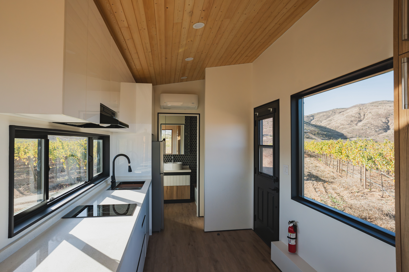 uxury tiny home kitchen with white quartz waterfall countertops, matte black fixtures, vaulted wood ceiling, large windows overlooking vineyard, and view through to hexagonal tile bathroom