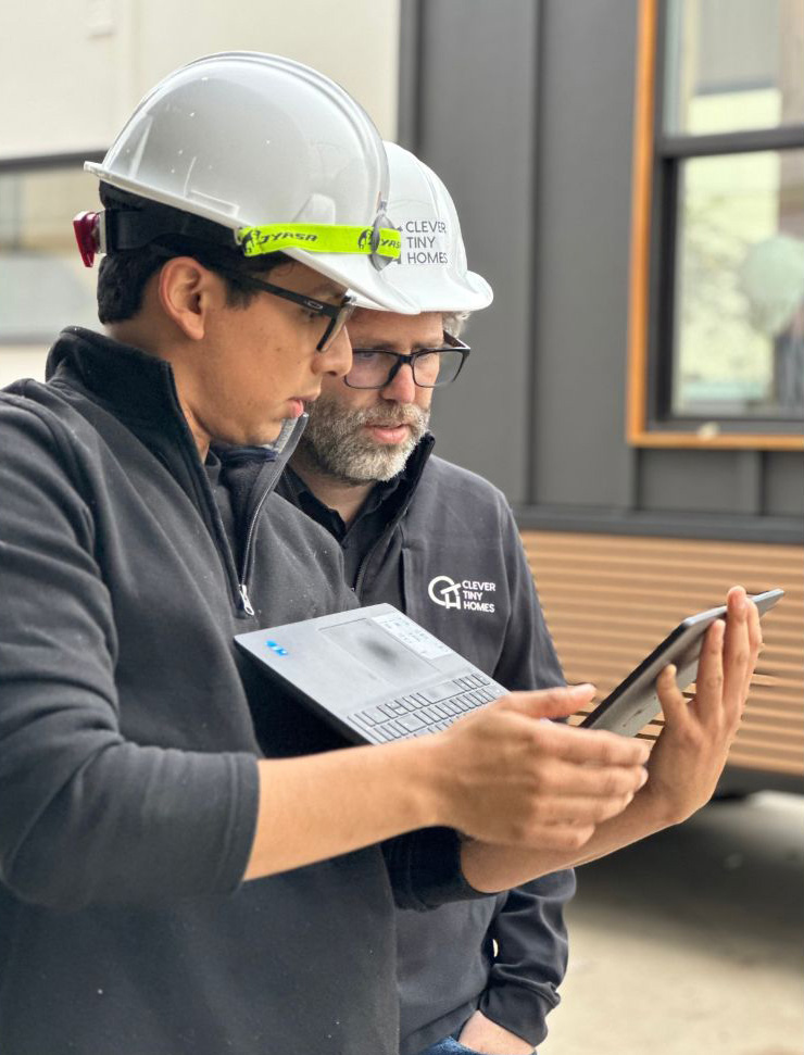 Two men in hard hats and safety gears are deep in conversation at a factory. A tiny home under construction is behind them, indicating an ongoing work on a project.