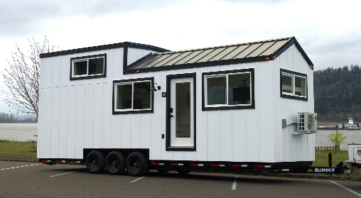 Exterior of a white Tiny Heirloom home on wheels with black trim, pitched metal roof, multiple windows, and a triple-axle trailer parked near a river and forested hills in Oregon.